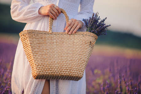 woman in white dress holding bouquet of lavender flowers in straw bag close upの写真素材