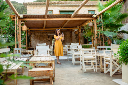 Woman in yellow dress walking by cafe terrace with smartphoneの写真素材