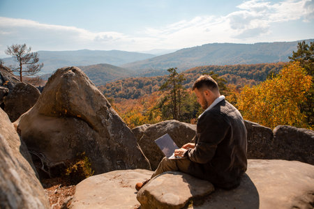 man freelancer working on laptop on the top of the rock with beautiful view of autumn forestの写真素材