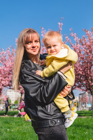 smiling mother holds her son by the hands in spring near pink cherry blossomsの写真素材