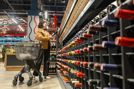 A female shopper carefully chooses a bottle of wine in a supermarket aisleの写真素材