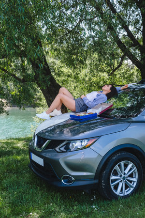 happy woman sitting on car hood eating pizza looking on river