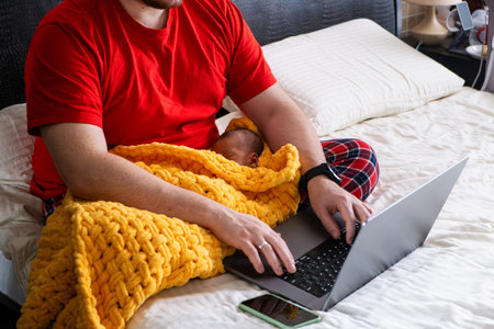 Man multitasking with laptop and baby on bedの写真素材