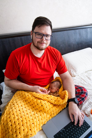 Man multitasking with laptop and baby on bedの写真素材