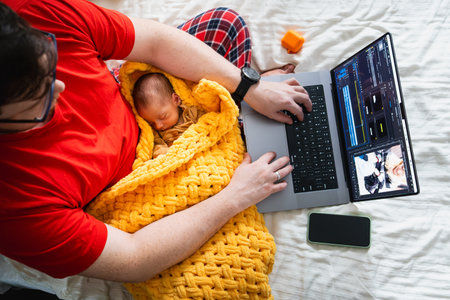 Man multitasking with laptop and baby on bedの写真素材