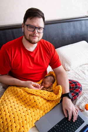 Man multitasking with laptop and baby on bedの写真素材