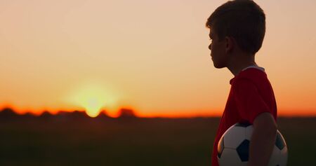 Young football player goes with the ball on the field dreaming of a football career, at sunset looking at the sunの写真素材