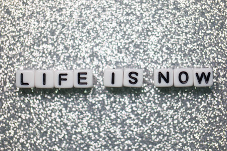 Black and white letter cubes spell out the phrase life is now on a shimmering silver surface, conveying a positive and motivational message about living in the present moment.の写真素材