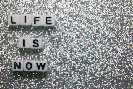 Words formed from square tiles spell out life is now, positioned against a shimmering backdrop. This artistic arrangement encourages mindfulness and appreciation of the present.の写真素材
