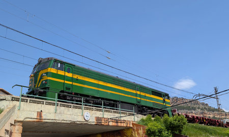 A vibrant green train with yellow stripes travels over an elevated track. It is set against a clear...