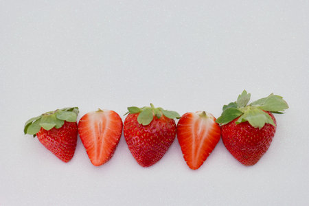 Five vibrant strawberries lined up on a light surface. Each berry showcases its red color and green leaves, highlighting their freshness and ripeness in natural light.の写真素材
