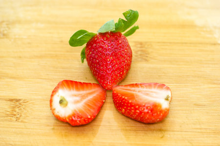 Juicy strawberries are sliced in half on a wooden cutting board, emphasizing their bright red flesh and green tops in a bright kitchen setting. The vibrant colors stand out in the clean environment.の写真素材