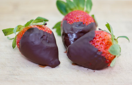 Three freshly dipped strawberries covered in smooth chocolate sit on a wooden surface, perfect for dessert at a summer gathering or romantic evening.の写真素材