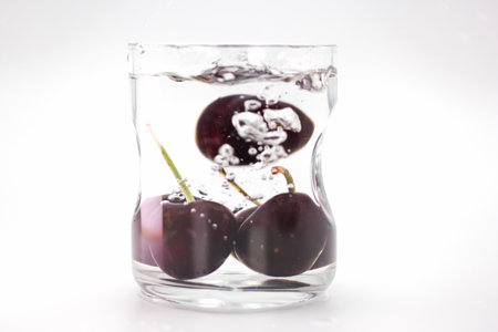 Fresh cherries are dropped into a glass of clear water, creating bubbles and a visually engaging moment as the fruit sinks and interacts with the liquid.の写真素材