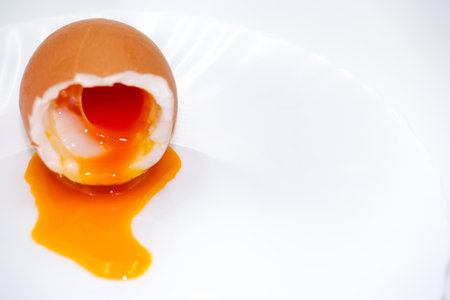 A soft boiled egg sits on a white plate, its shell cracked open to reveal a perfectly cooked yolk oozing out. Perfect for a nutritious breakfast or brunch option.の写真素材