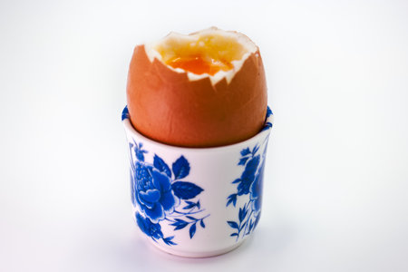 A soft boiled egg sits in a beautifully designed blue and white cup. The top of the egg is cracked open, revealing the golden yolk inside. This delicious breakfast features an elegant presentation.の写真素材