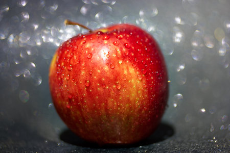 A vibrant red apple covered in glistening water droplets sits against a dark background that reflects soft light, highlighting its fresh appearance and texture.の写真素材