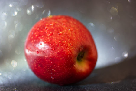 A vibrant red apple rests on a dark surface, covered in tiny water droplets that glisten in the light. The blurred backdrop creates a dreamy ambiance, emphasizing the fruit's freshness.の写真素材