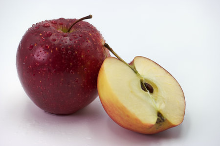 A vibrant red apple rests beside a neatly sliced half, showing its juicy interior and seeds. Water droplets enhance the fresh appearance against a bright background.の写真素材