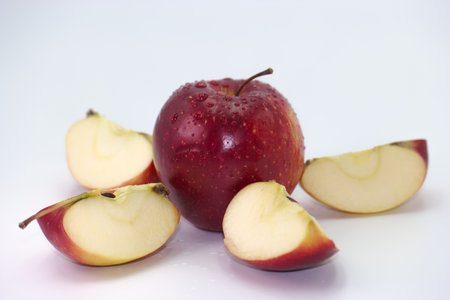 A bright red apple sits intact while several slices of the fruit are arranged nearby on a smooth white surface. The apple is glistening with moisture, indicating freshness.の写真素材