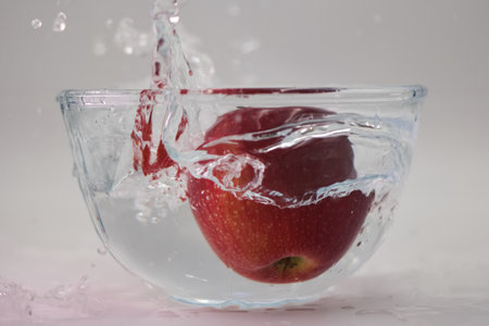 A vibrant red apple drops into a bowl filled with clear water, creating an exciting splash. Water droplets scatter around, showing the freshness of the fruit in a cozy kitchen setting.の写真素材