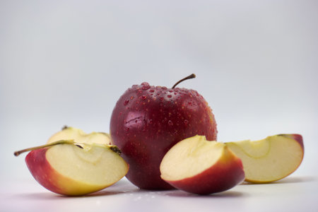 A shiny red apple is surrounded by freshly cut slices, showing a mix of vibrant colors and textures. Dew droplets glisten on the surface, adding freshness to the display.の写真素材