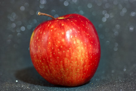 A vibrant red apple is positioned on a dark surface that sparkles under soft lighting. The shiny skin of the apple reflects light, showing its fresh appearance and delightful color.の写真素材