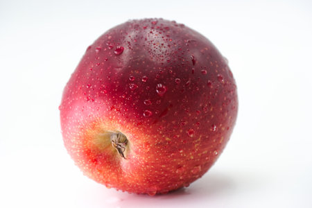A vibrant red apple is placed on a smooth white surface, glistening with water droplets that emphasize its freshness and appealing texture. It stands out due to its vivid colors and detail.の写真素材