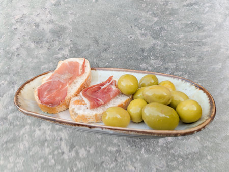 A platter holds two slices of bread spread with creamy topping and garnished with savory cured meat next to green olives. This snack reflects Mediterranean culinary traditions.の写真素材