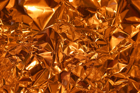 Shiny, crinkled orange foil displays a rich texture with varying light reflections. The intricate folds create depth and visual interest, evoking warmth and energy.の写真素材