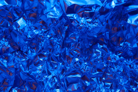Crumpled blue aluminum foil covers the surface, creating a vibrant, shiny texture. The light reflects off the metallic surface, enhancing its blue hue and intricate folds.の写真素材
