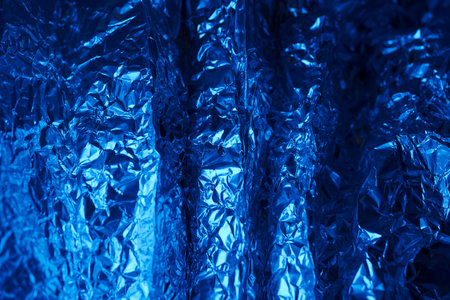 Crumpled aluminum foil in a deep blue hue creates an eye catching texture, accentuated by light that highlights the creases and folds, emphasizing its metallic quality.の写真素材