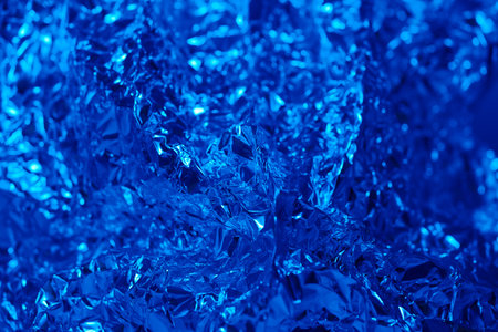 Textured blue foil with glossy surface reflects light, creating a vibrant abstract design. The intricately wrinkles and folds showcase depth and motion, offering a rich visual experience.の写真素材