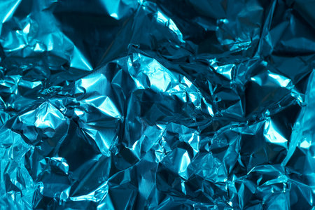 Crumpled blue foil creates a dynamic surface that catches and reflects light, showing various abstract textures and shapes. The shimmering surface adds depth to the overall effect.の写真素材