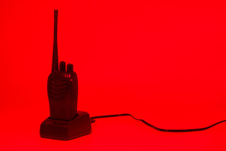 A black walkie talkie stands on a simple base against a vibrant red backdrop. The device is...