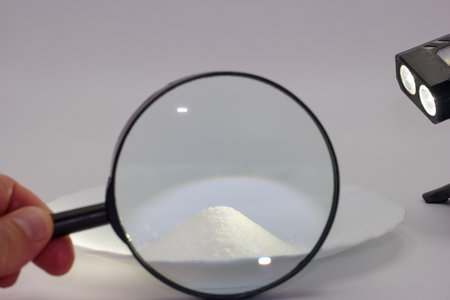 A hand holds a magnifying glass focused on a small pile of white powdered substance, illuminated by...
