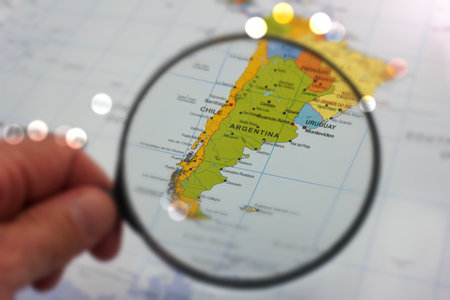 A hand holds a magnifying glass above a map of South America, highlighting the countries of...