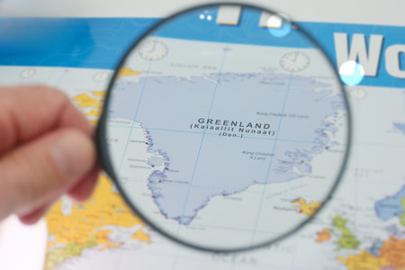 A hand holds a magnifying glass over a map of Greenland highlighting its position within the Arctic...