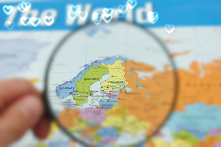 A hand holds a magnifying glass over a map that highlights Finland and Sweden in northern Europe,...