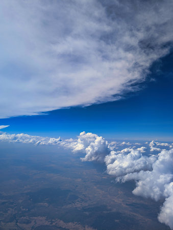Bright sky with varied cloud formations and a deep blue gradient observed from an airplane at cruising altitude. The landscape below adds depth to the atmospheric view.の写真素材