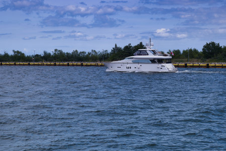 A sleek luxury yacht cruises smoothly across calm waters near a harbor, surrounded by green trees under a bright sky and occasional clouds in the afternoon light.の写真素材