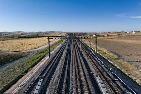 Long stretches of train tracks extend through an open rural area, surrounded by fields and distant hills. A vibrant blue sky enhances the serene atmosphere in this midday view.の写真素材