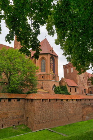 Beautiful medieval castle made of red brick is framed by lush green trees. Intricate architectural...