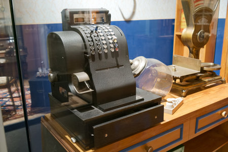 A vintage cash register sits behind glass in a museum, showing the mechanical design and style from...