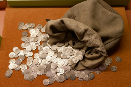 A cloth bag lies open with numerous silver coins scattered around it, showing a historical...