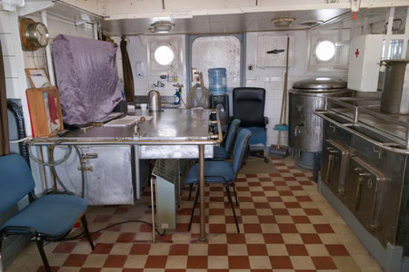 Inside a ship's kitchen, stainless steel counters and cooking equipment are visible. Blue chairs...