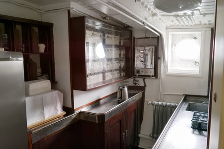 Inside a ship's kitchen, stainless steel surfaces contrast with wooden cabinets. Shelves show...
