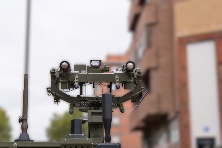 A military surveillance device is positioned on a street in a city. It features two large cameras and various sensors, overlooking nearby buildings under a cloudy sky.の写真素材