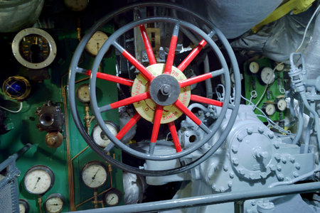 A close up of a submarine's control room features a red steering wheel surrounded by numerous gauges...