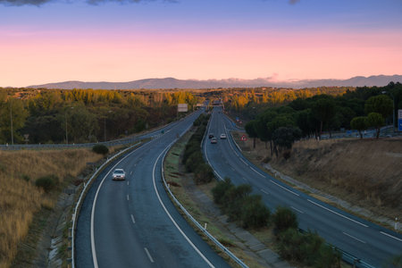 Two parallel highways stretch into the distance under a colorful sunset sky. Lush trees line the roads, and cars are visible on the empty lanes, creating a peaceful atmosphere.の写真素材